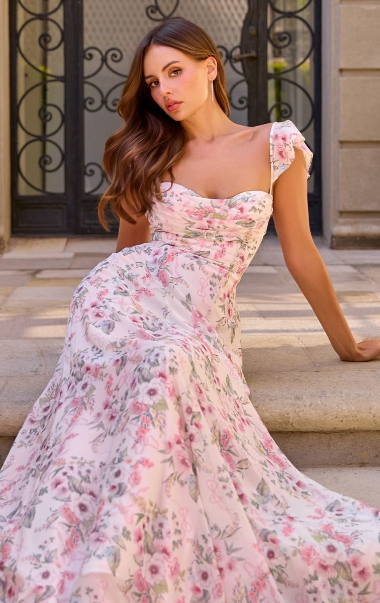 A woman wearing a flowing floral gown with puff sleeves and a sweetheart neckline, posed on stone steps in front of an ornate gate.