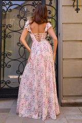 A woman wearing a pink floral maxi dress with a lace-up open back, photographed from behind in front of a stone wall and black wrought iron gate.