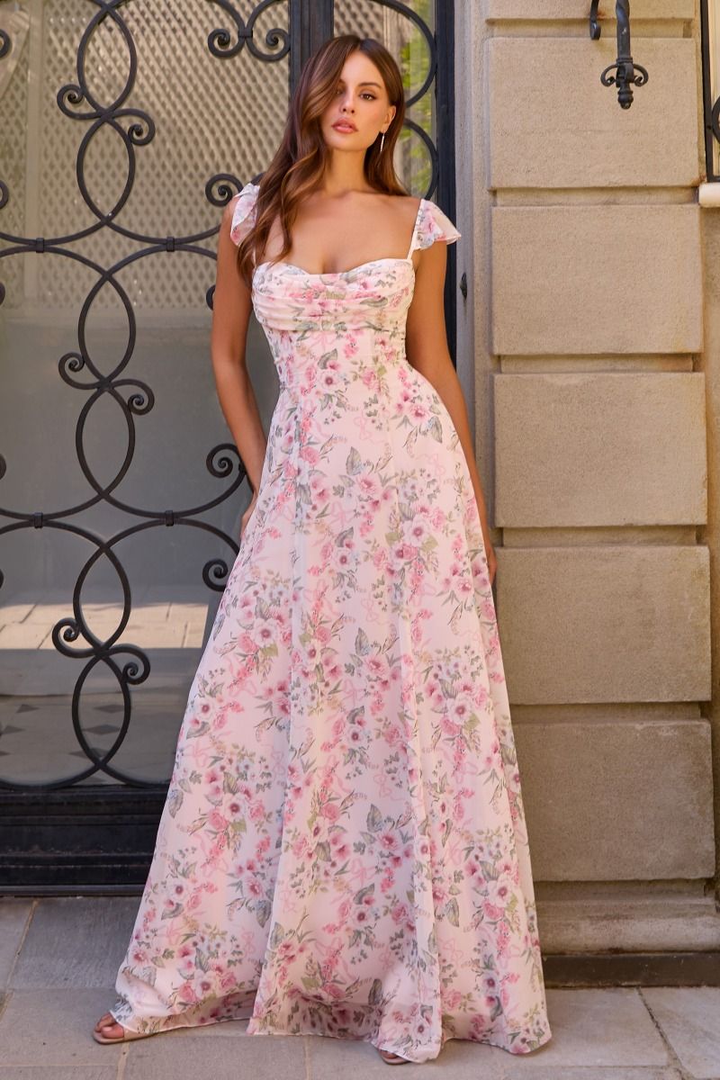A woman wearing a floor-length floral maxi dress with a sweetheart neckline and cap sleeves, standing outdoors in front of a stone wall and black wrought iron gate.