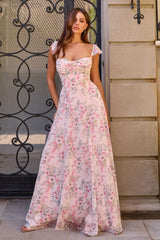 A woman wearing a floor-length floral maxi dress with a sweetheart neckline and cap sleeves, standing outdoors in front of a stone wall and black wrought iron gate.