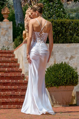 Woman in a white evening gown standing outdoors with greenery and a stone wall in the background