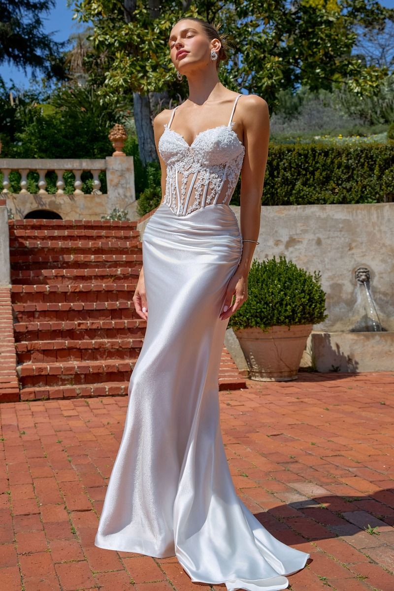 Woman in a white wedding dress standing outdoors with greenery and a fountain in the background